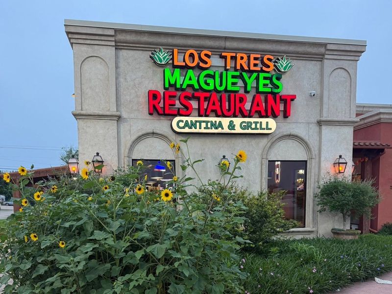 The lit sign and entrance of Los Tres Magueyes Restaurant Cantina and Grill at dusk.