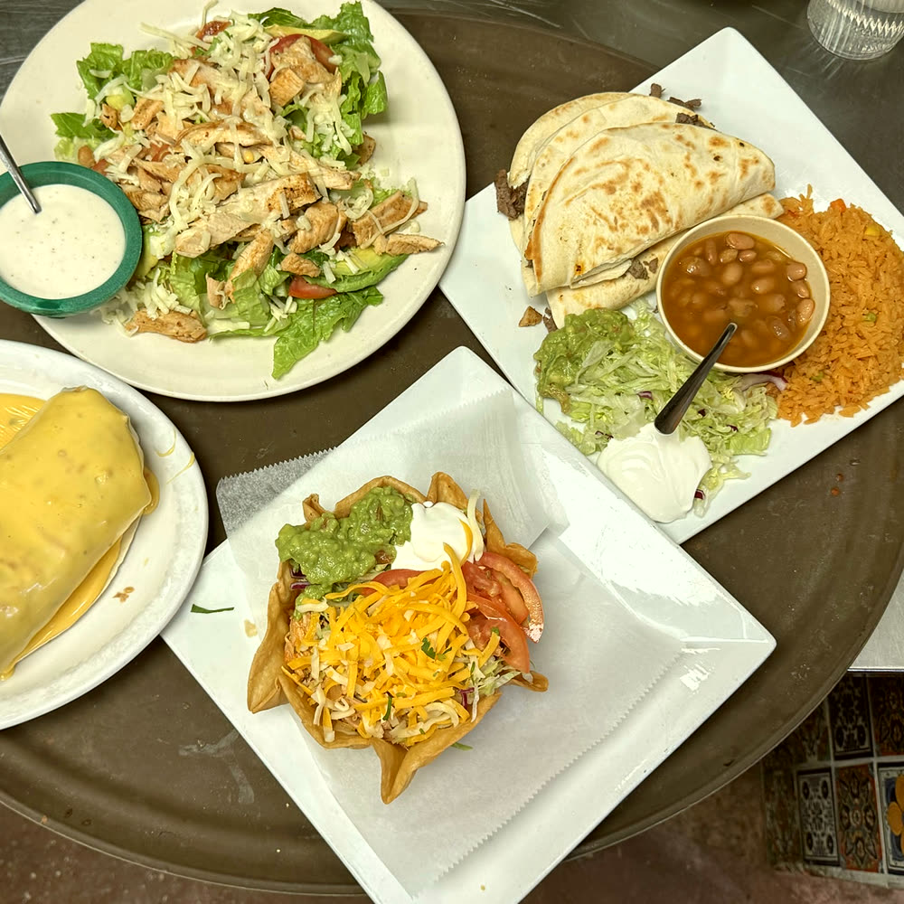 Four plates on a serving tray: a grilled chicken salad, a taco salad bowl, quesadillas with rice and charro beans, and a plate smothered in chile con queso.