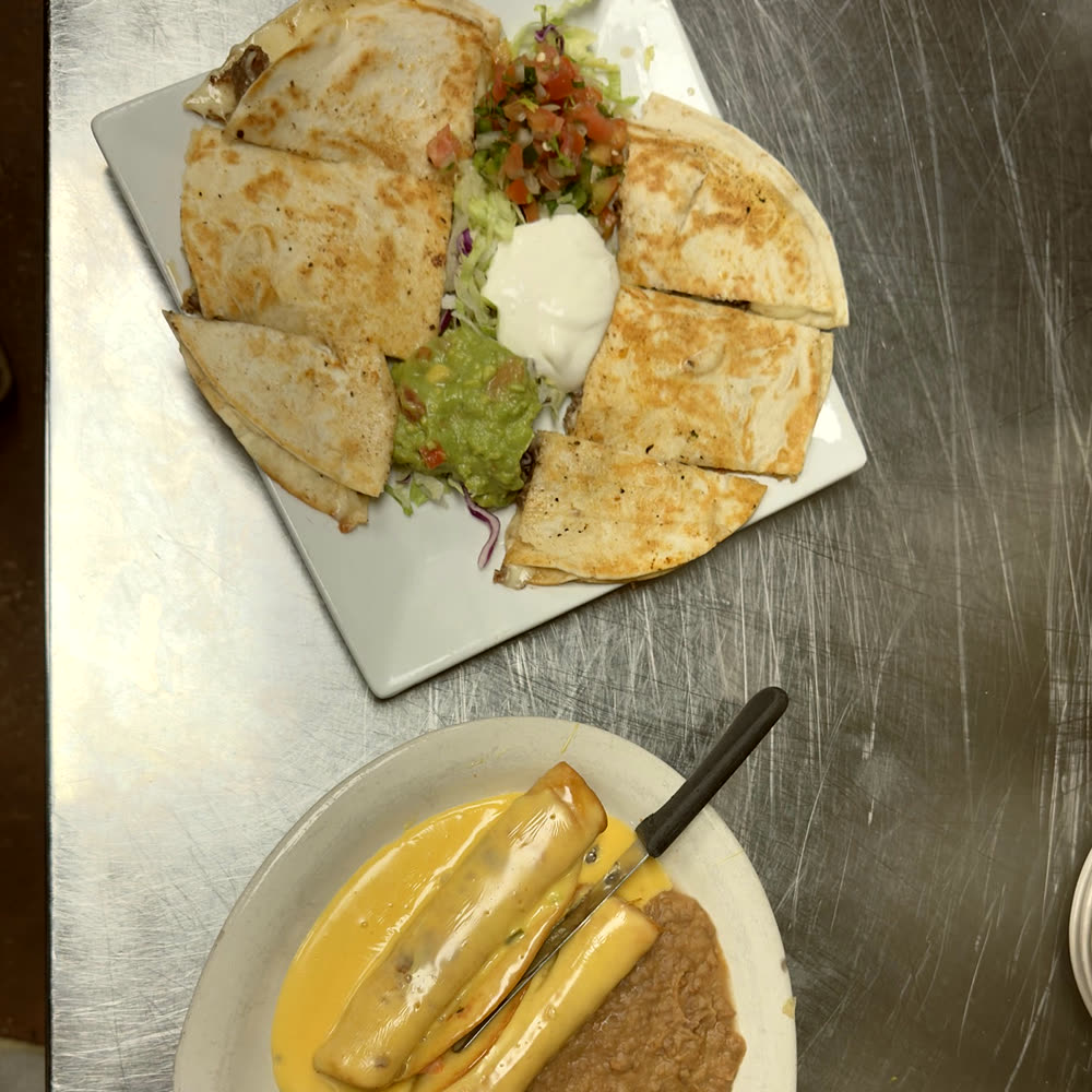 Quesadillas cut into wedges around guacamole, sour cream, pico de gallo and lettuce, beside a plate smothered in chile con queso with refried beans.