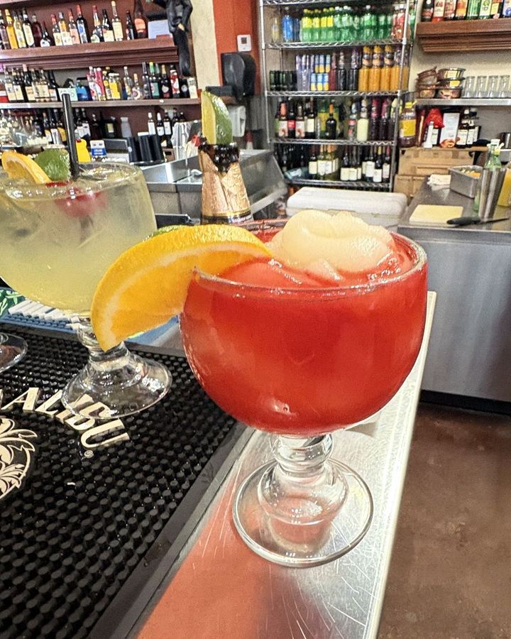 A red strawberry margarita over ice in a big goblet, topped with a frozen scoop and an orange slice, a lime margarita beside it on the bar.