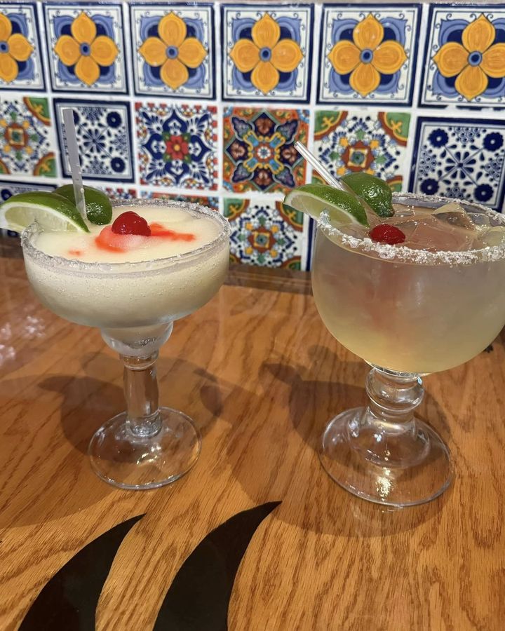 Two margaritas, one frozen and one on the rocks, each with lime and a cherry, on a wooden table in front of a hand-painted talavera tile wall.