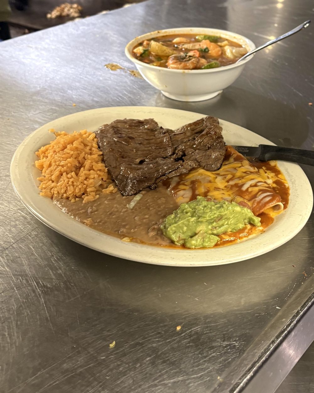 Grilled steak with enchiladas in red sauce and melted cheese, rice, refried beans and guacamole on the steel counter of the kitchen pass, a bowl of shrimp soup behind it.