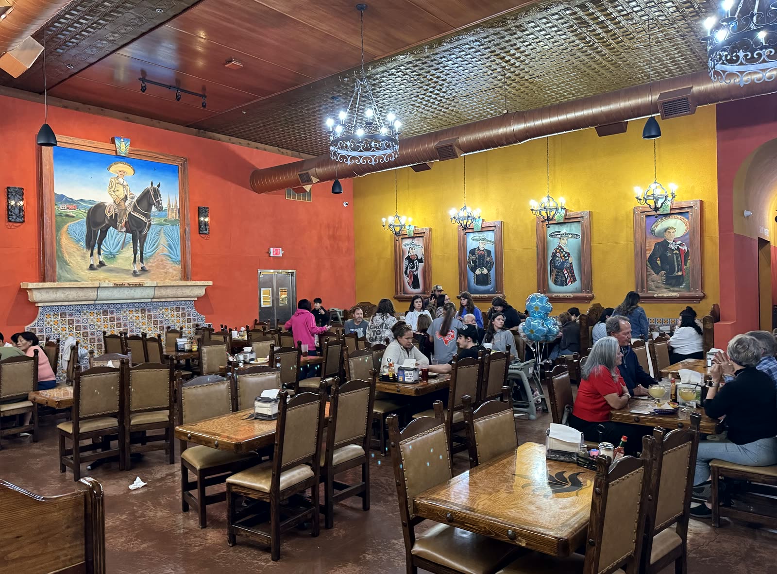 The main dining room: orange and marigold walls, the Vicente Fernandez mural, charro portraits and carved wooden chairs.